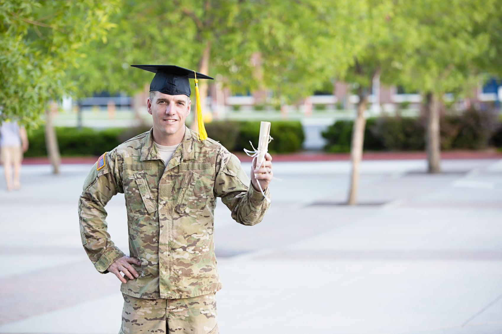 A service member in fatigues wearing a graduation cap holding a diploma.